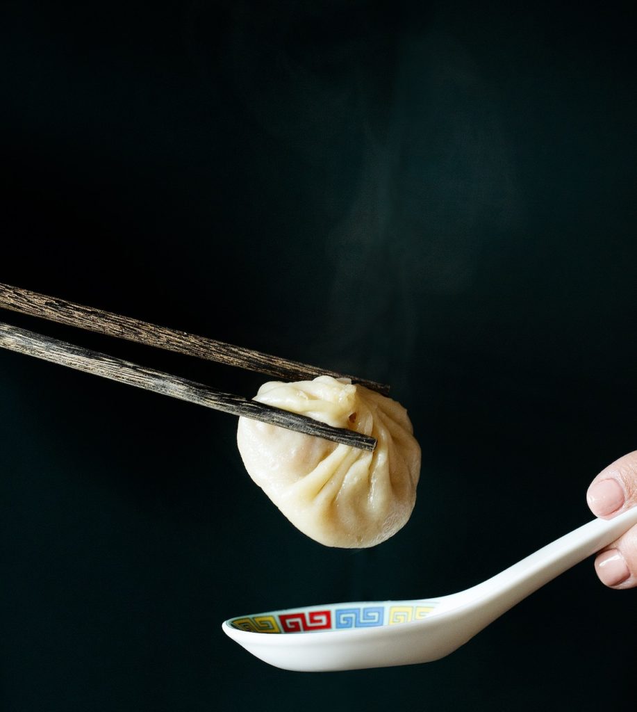 Image of a pair of chopsticks floating in from the left, holding a soup dumpling over a Chinese soup spoon floating in from the right. The background is dark.