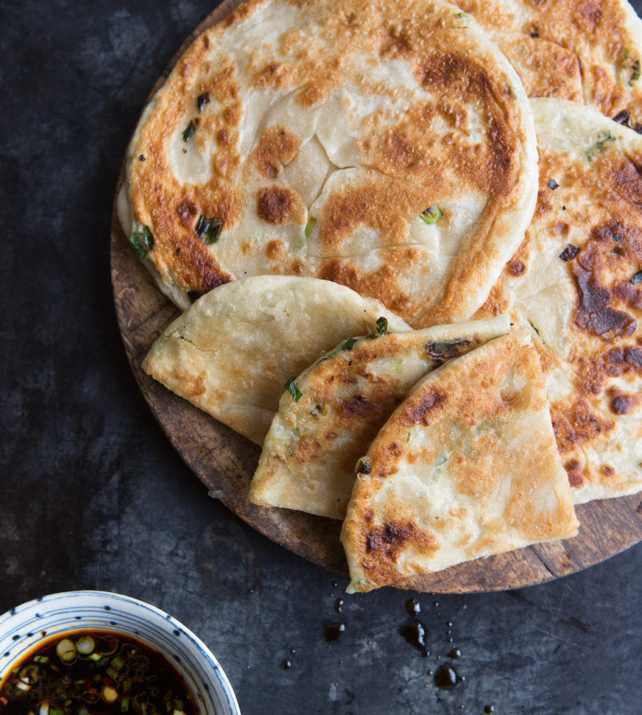 Green Onion Pancakes from Chinese Soul Food cookbook by Hsiao-Ching Chou. Several pancakes are arranged on a wooden plate and the plate sits on a textured dark background. Photo credit: Clare Barboza