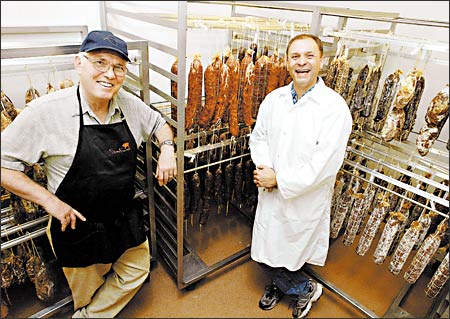 Image of Armandino Batali and his son-in-law Brian D'Amato. They have racks of curing salami behind them.